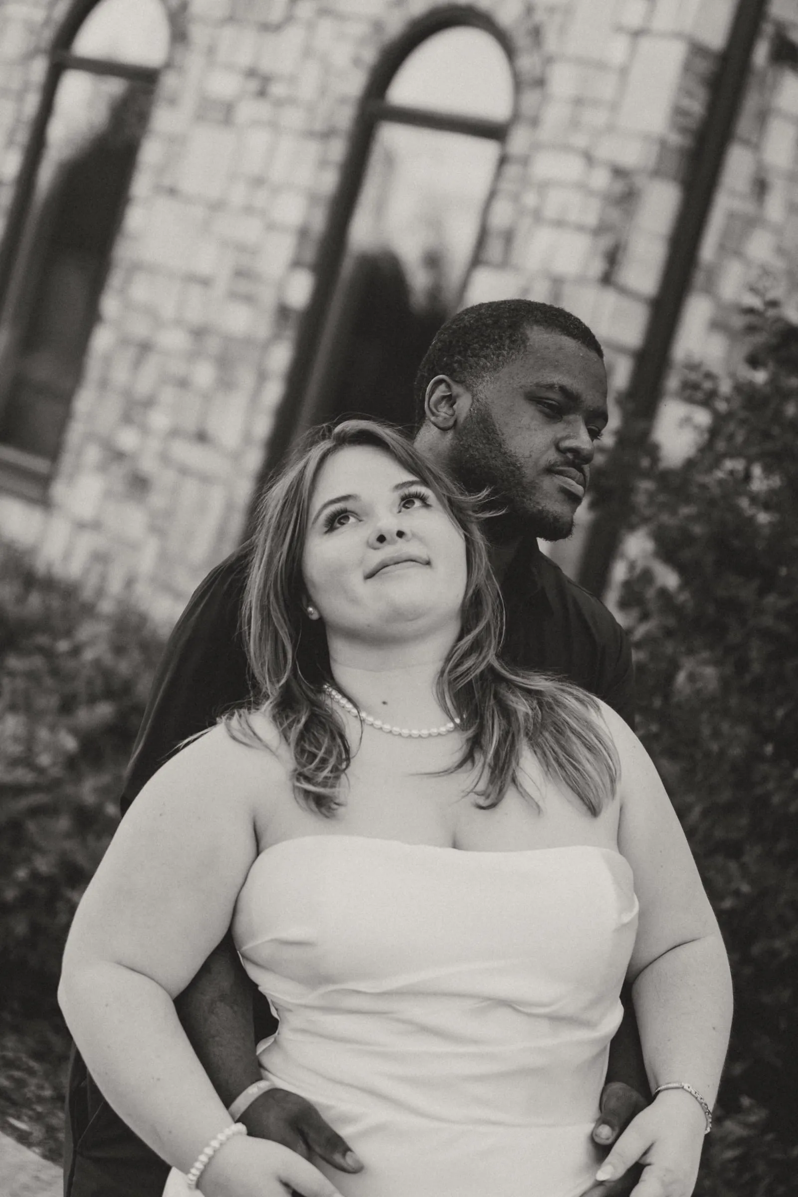 A bride smiling as her groom holds her close in front of a stone chapel, in black and white