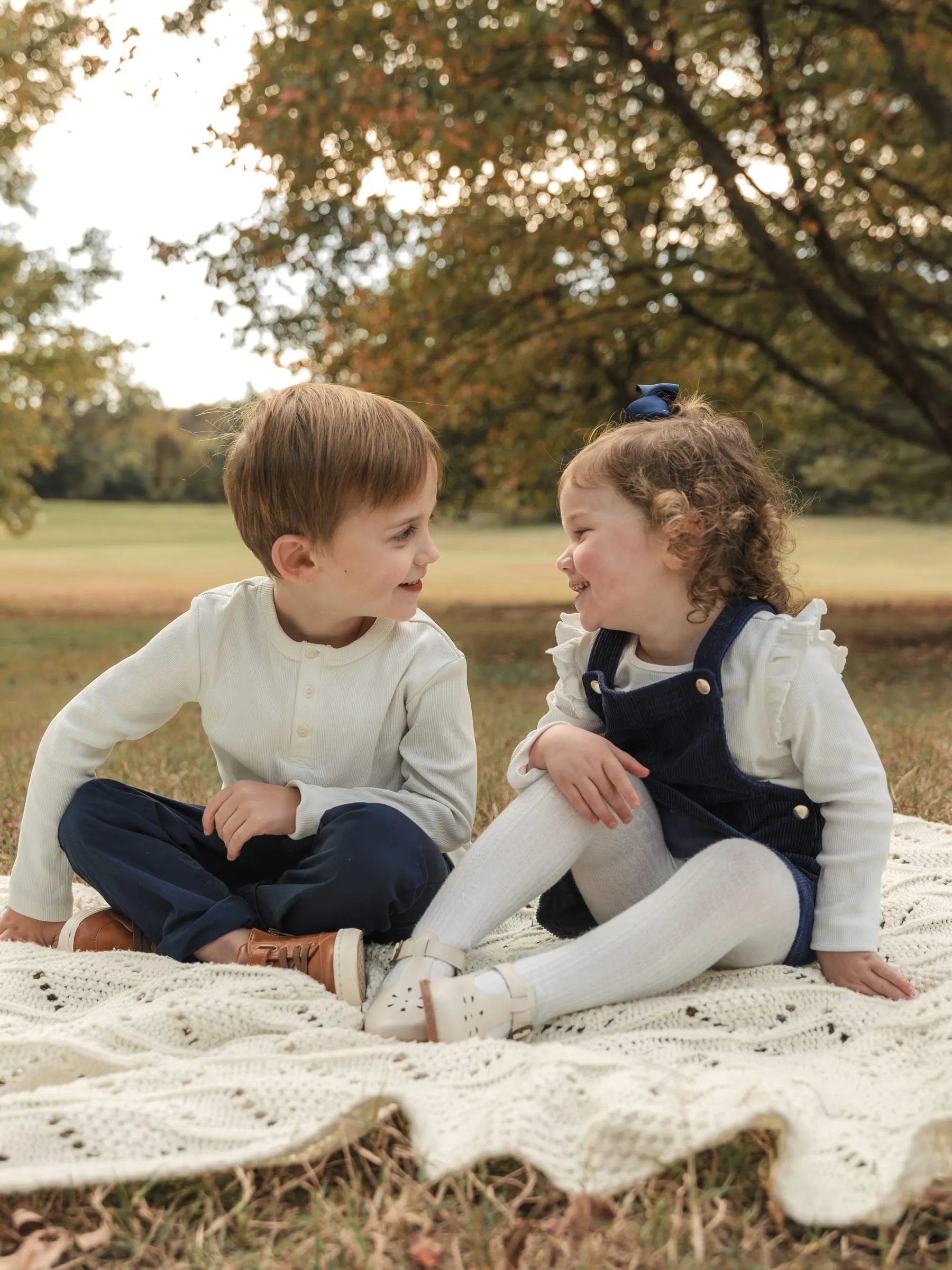 Two little ones sitting on a cream blanket in a sunny field, smiling at each other