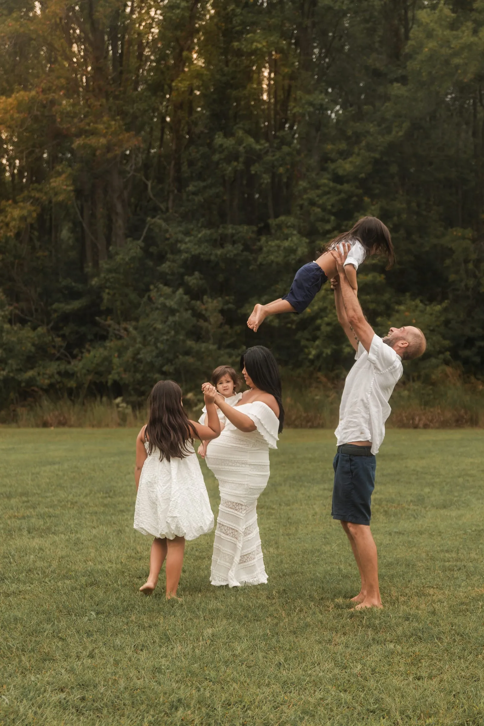 Dad tossing his daughter into the air while an expecting mom and big sister watch in a field