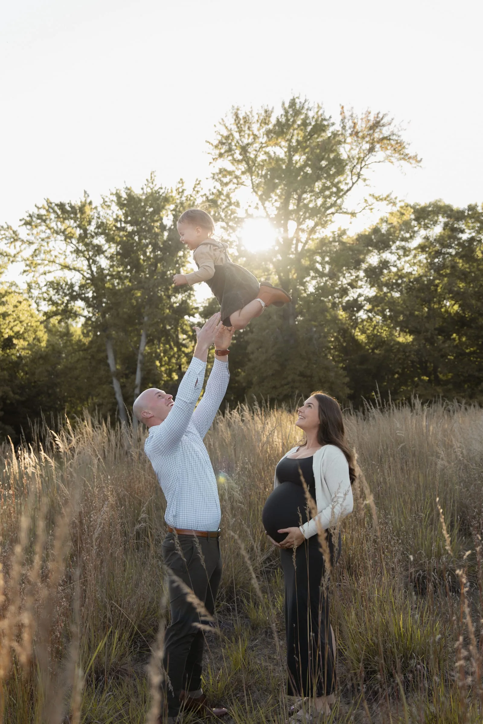 A dad tossing his little girl into the sunset sky beside his pregnant wife in a golden field