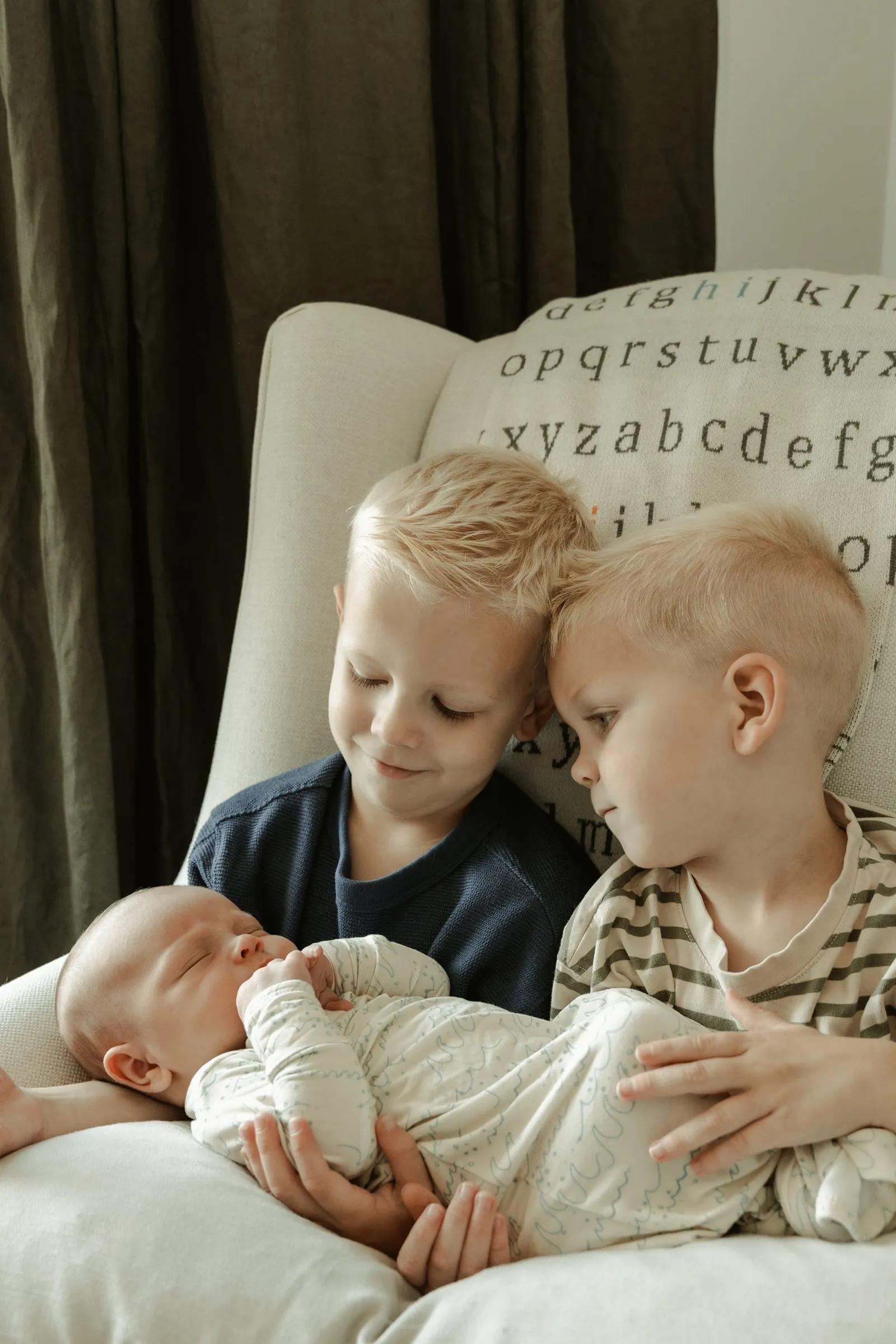 Two big brothers snuggling their newborn sibling in a nursery armchair