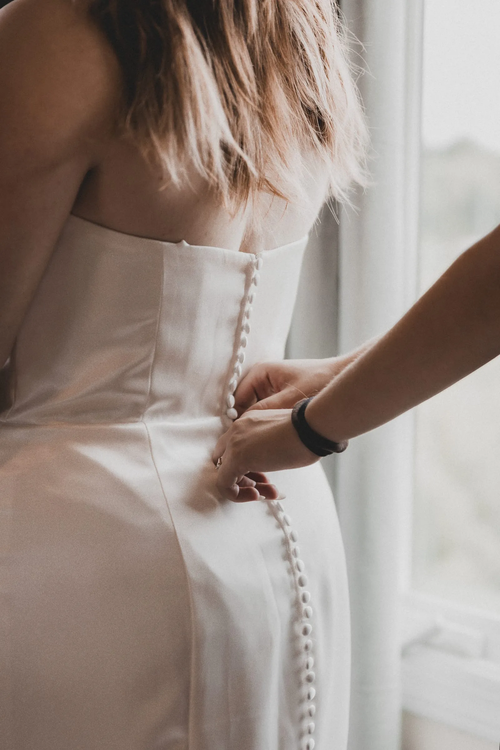 The last buttons of a wedding gown being fastened by the window
