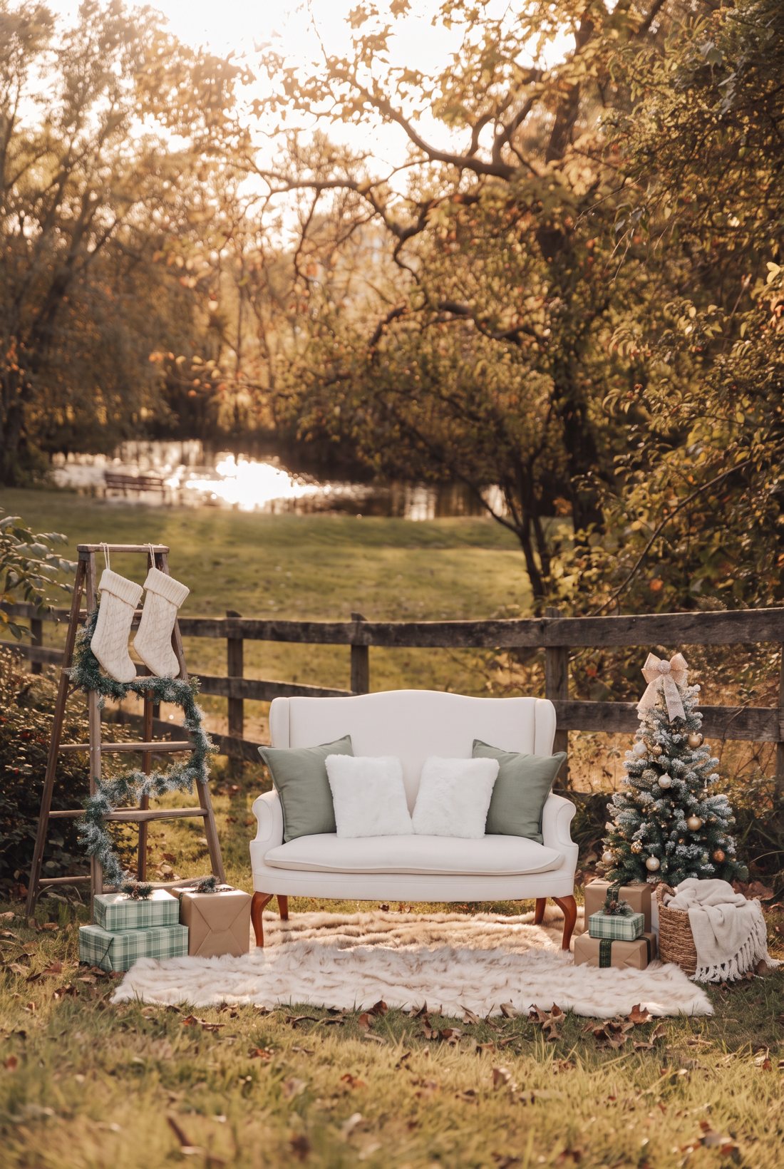 The holiday setup: a white wingback loveseat on a cream fur rug with sage and white pillows, a ladder with stockings and garland, a small flocked tree and wrapped gifts, in front of a wooden fence at the park